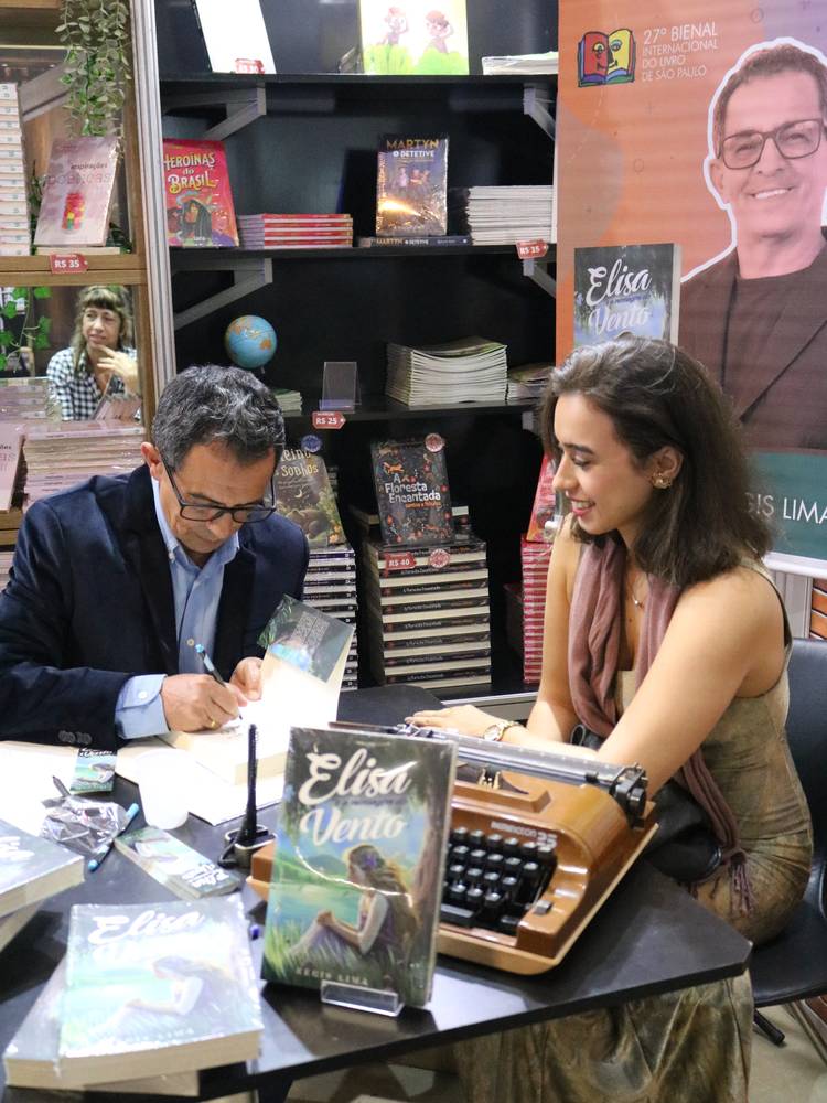 Régis Lima sozinho à mesa de autógrafos, caneta na mão, assinando um exemplar